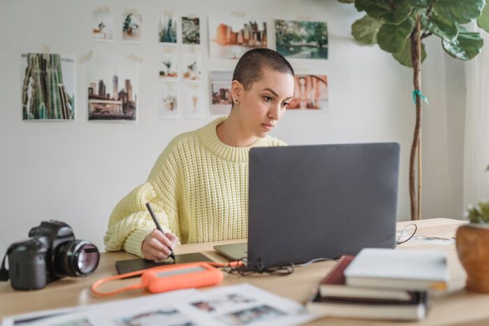 Person in a yellow sweater working on a laptop at a desk with a camera and photos, highlighting potential job impacts.