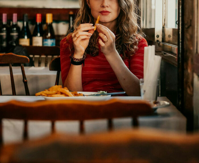 Woman in a striped shirt holding fries, demonstrating a frugal choice in a cozy restaurant setting with wine bottles in the background.