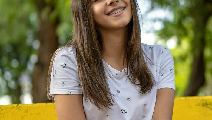 Smiling girl with braces in a white shirt sitting on a yellow bench outdoors.