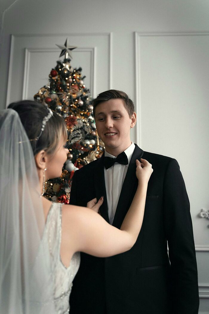 Bride adjusting groom's bow tie in front of a decorated Christmas tree, highlighting wedding guest expectations.