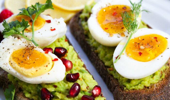 Avocado toast with boiled eggs, garnished with pomegranate seeds and herbs, on a white plate.