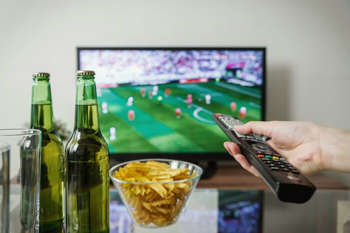 A person uses a remote to watch soccer on TV with beer and snacks, reminiscent of weird government projects.