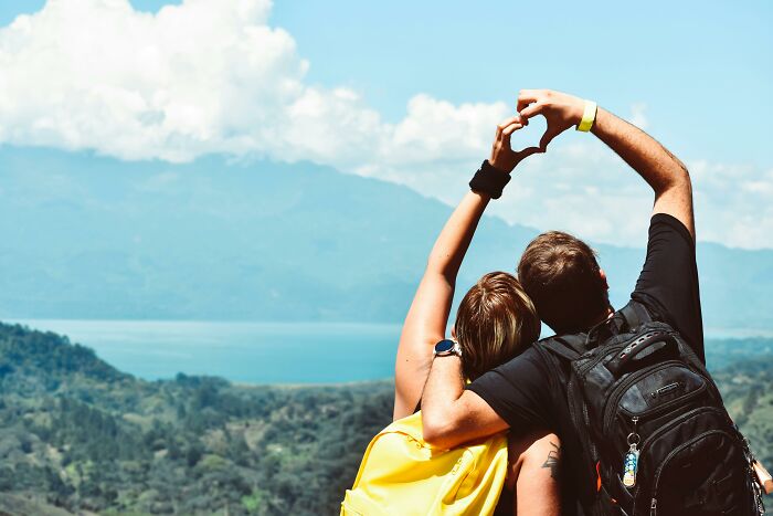 Adults forming a heart shape with their hands, facing a scenic mountain and lake view, symbolizing harsh life lessons in nature.