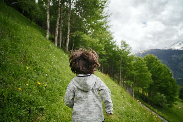 Child in a gray hoodie running through a lush green field surrounded by trees and mountains, capturing a sense of freedom.