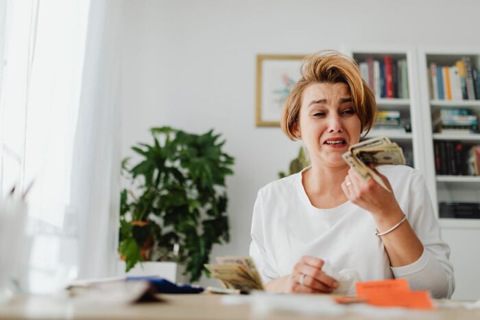 Person in a white shirt looking distressed while holding money, with a plant and bookshelf in the background. Ringtones theme implied.