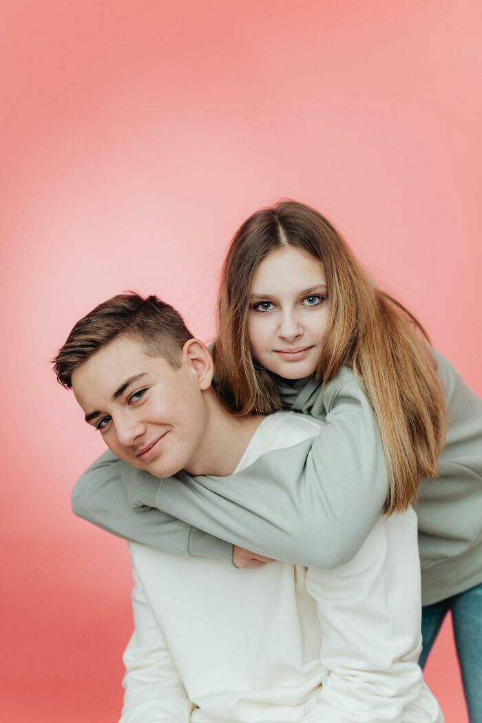 Two siblings smiling together, posing playfully with one giving a piggyback ride against a pink background.
