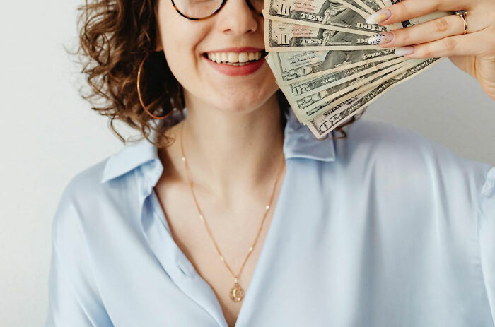Happy woman in a blue shirt holding a fan of dollar bills, illustrating frugal living.