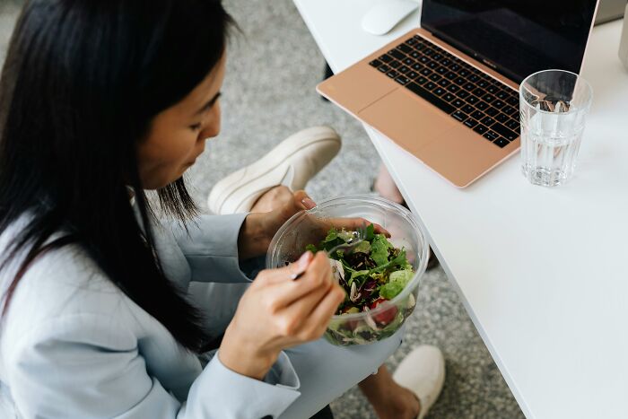 Person eating a salad at a desk, dealing with work-struggles, next to a laptop and a glass of water.