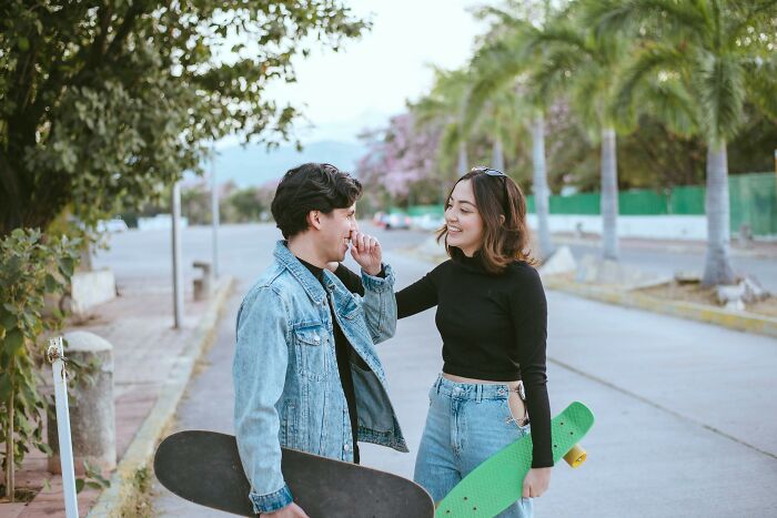 A couple smiling and holding skateboards on a tree-lined street, showing love and companionship.