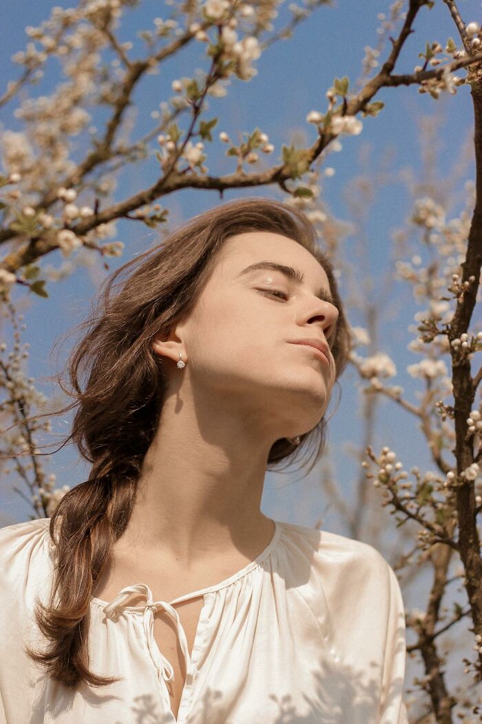Person with prominent nose enjoying sunlight among flowering branches, showcasing unconventional beauty.