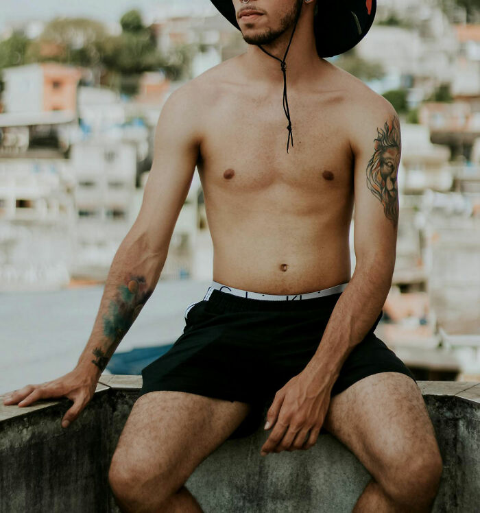Shirtless person with tattoos on each arm, wearing a hat and shorts, sitting on a wall, showcasing beauty in unconventional features.