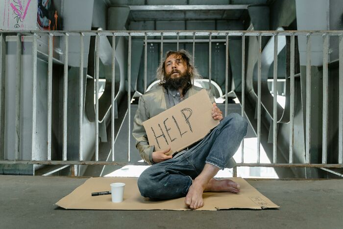 Homeless man under a bridge holding a "HELP" sign, representing harsh realities about life.