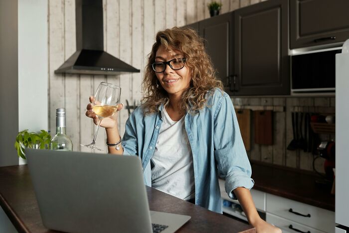 Woman with glasses in a kitchen, holding a wine glass, watching a laptop, possibly exploring weird government projects online.