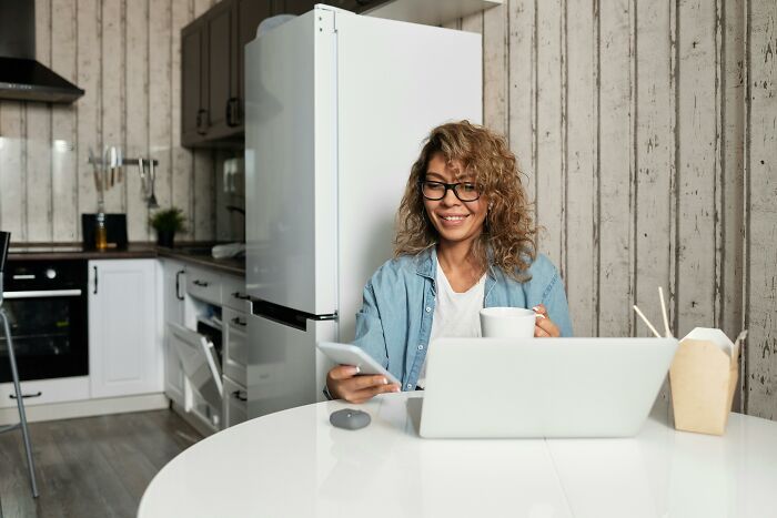Woman enjoying perks of not having to go to the office, working remotely with laptop and phone in a cozy kitchen setting.