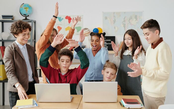 Students gathered excitedly around laptops, participating in a weird government project activity in a classroom.