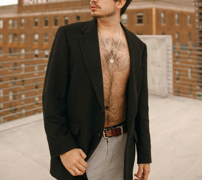 Man standing on rooftop in a black blazer with prominent nose, showcasing unconventional beauty against an urban background.