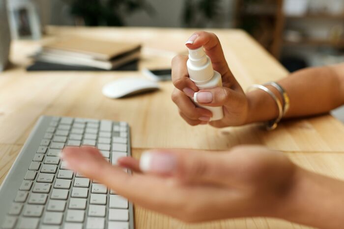 Person using multipurpose spray near a keyboard on a wooden desk.