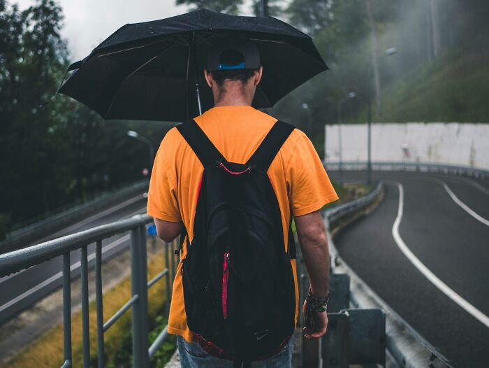 Man wearing orange shirt and backpack holding umbrella while walking alone on rainy road, reflecting on real men myths.