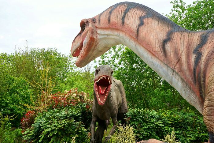 Two dinosaur statues in a garden setting, appearing as though in a hilarious overheard conversation.