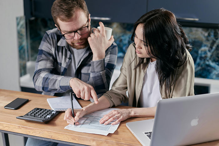 A couple discussing finances over a laptop, calculator, and documents at a kitchen table. A couple discussing finances over a laptop, calculator, and documents at a kitchen table.
