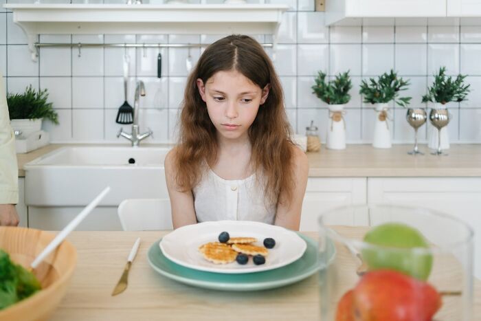 Young girl at a kitchen table, looking at pancakes with blueberries, appears thoughtful or concerned.