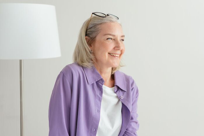 Woman with a prominent nose smiling, wearing glasses and a purple shirt, standing near a lamp in a bright room.