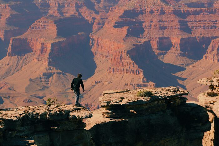 Person standing on the edge of a cliff, overlooking a vast natural wonder of expansive canyons and rugged terrain.
