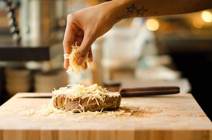 A hand sprinkling cheese over a slice of bread on a wooden board, demonstrating a cooking cheat.