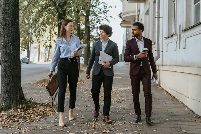 Three people walking on a sidewalk in business attire, holding papers and coffee, sharing life cheat codes.