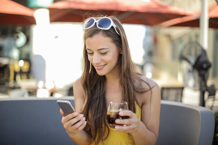 Woman smiling at phone, outdoors with a drink, reflecting on songs as ringtones.