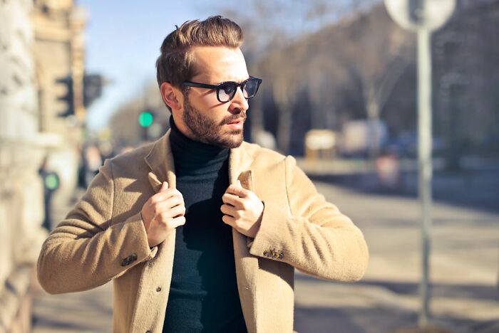Man in sunglasses and a camel coat strolling confidently outdoors, addressing perceptions of men's actions and judgments.