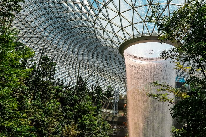 Indoor waterfall in a glass-domed structure surrounded by lush greenery, highlighting cultural differences in architecture.