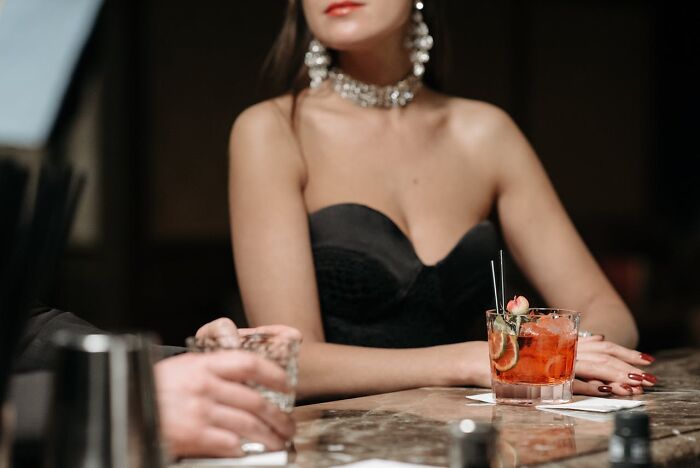 Woman in elegant attire at a bar, holding a cocktail, highlighting issues wedding guests dislike.