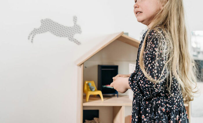 Young girl standing near a dollhouse, wearing a floral dress, in a room with a rabbit decal on the wall.