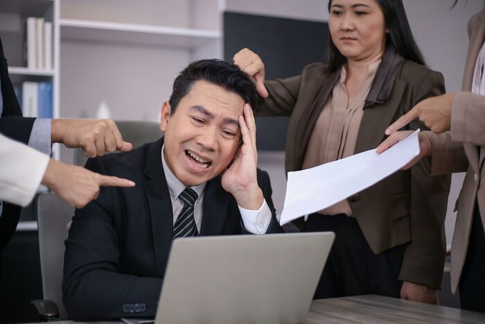 Stressed man at a laptop, surrounded by colleagues pointing fingers, illustrating harsh realities about life.