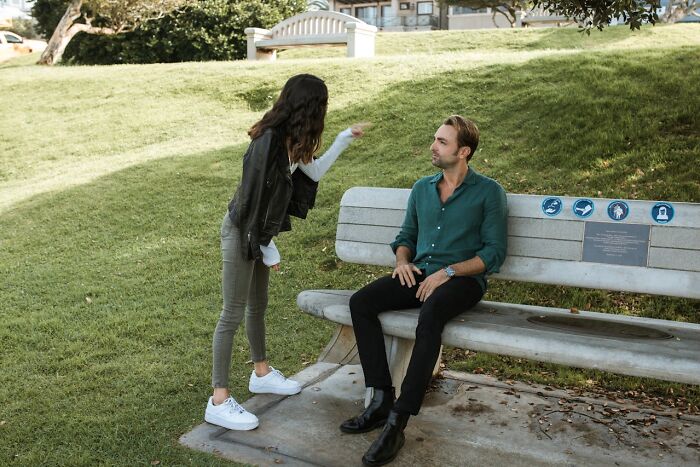 A woman gestures while a man sits on a park bench, discussing cheap versus frugal living.