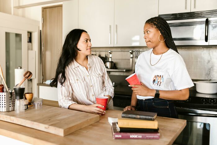 Two women in a kitchen sharing a funny conversation over red cups, capturing hilarious overheard conversations.