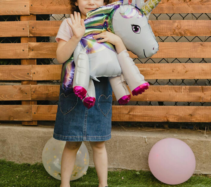Child holding a unicorn balloon, standing in front of a wooden fence with balloons nearby.
