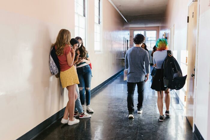 Teens in a school hallway, chatting and walking, possibly sharing hilarious overheard conversations.