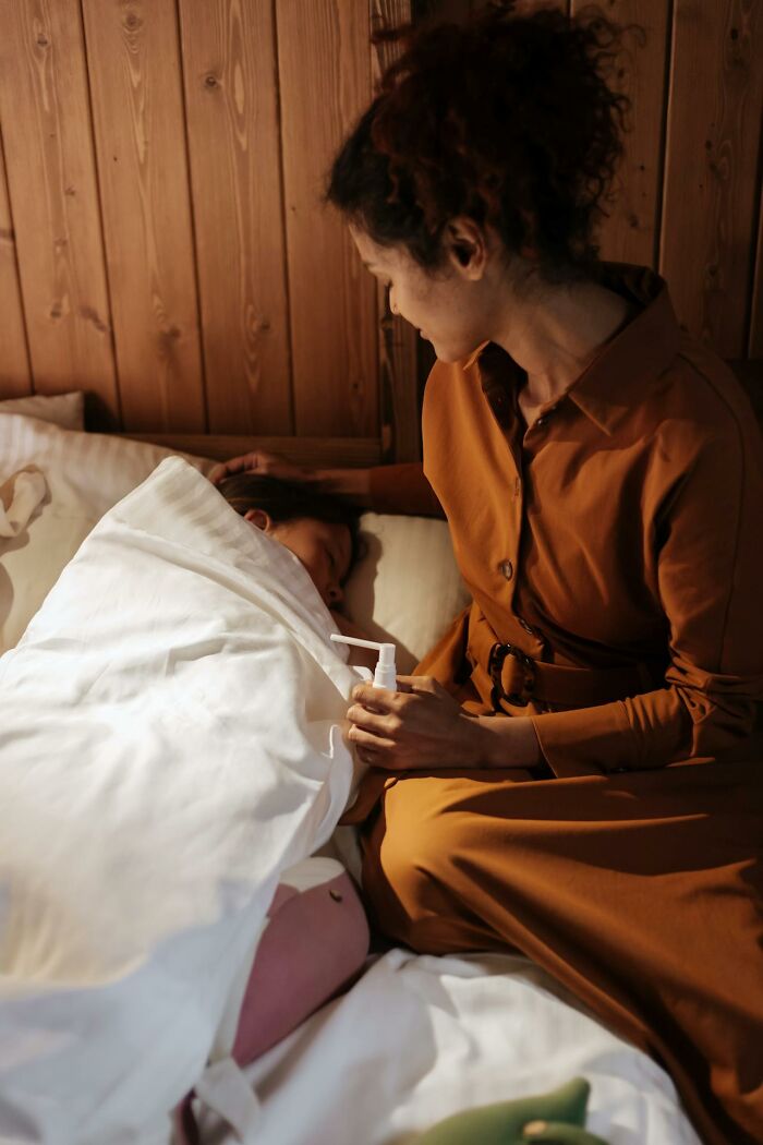 A woman in a brown dress attends to a child sleeping in bed, holding a white spray bottle, in a cozy wooden room.