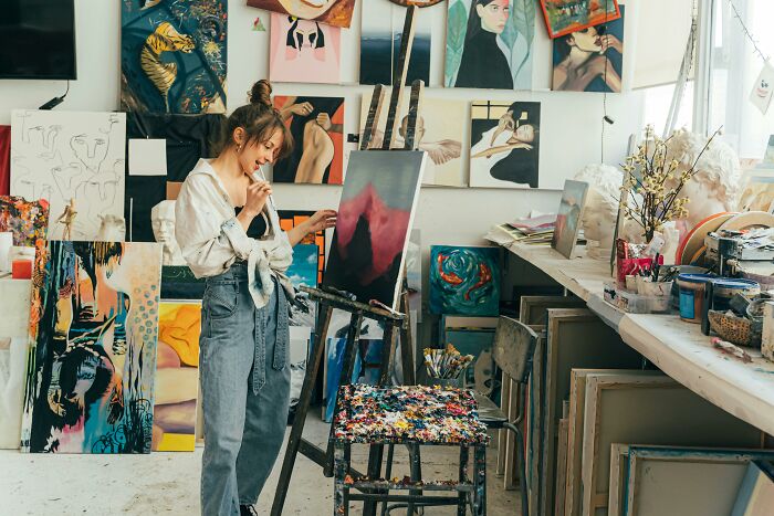 Artist contemplating life, surrounded by paintings in a cluttered studio, reflecting harsh realities through her art.