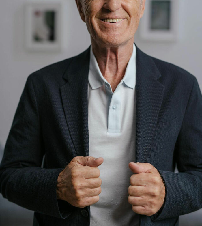Elderly man smiling, adjusting his jacket in a dimly lit room, possibly sharing a creepy family story.