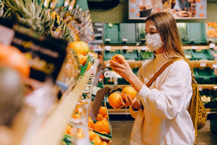 Person in a grocery store wearing a mask, selecting oranges, illustrating frugal shopping habits.