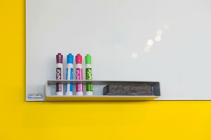 Whiteboard with markers and eraser on tray, set against a vibrant yellow wall, illustrating organization in everyday life.