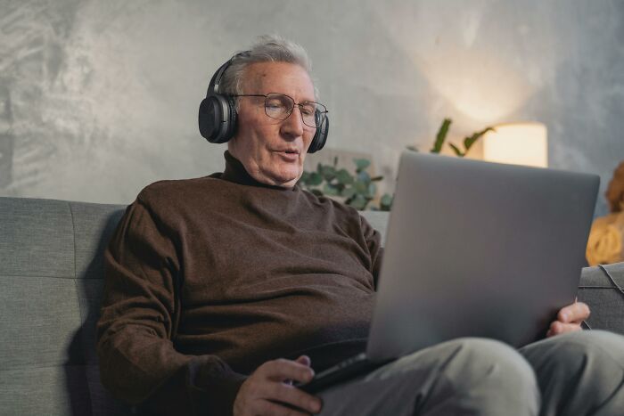 Elderly man experiencing eye-opening moments, wearing headphones, focused on a laptop in a cozy setting.
