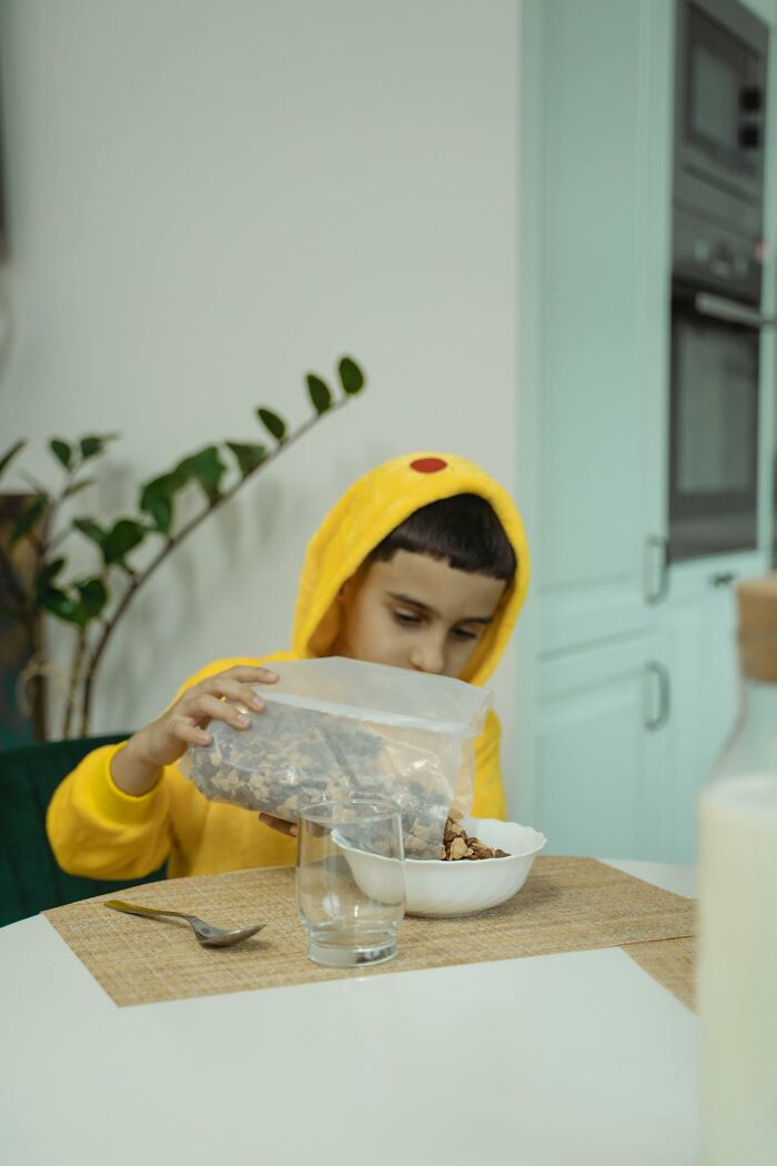 Child in yellow hoodie, practicing frugal habits by pouring cereal into a bowl at a kitchen table.