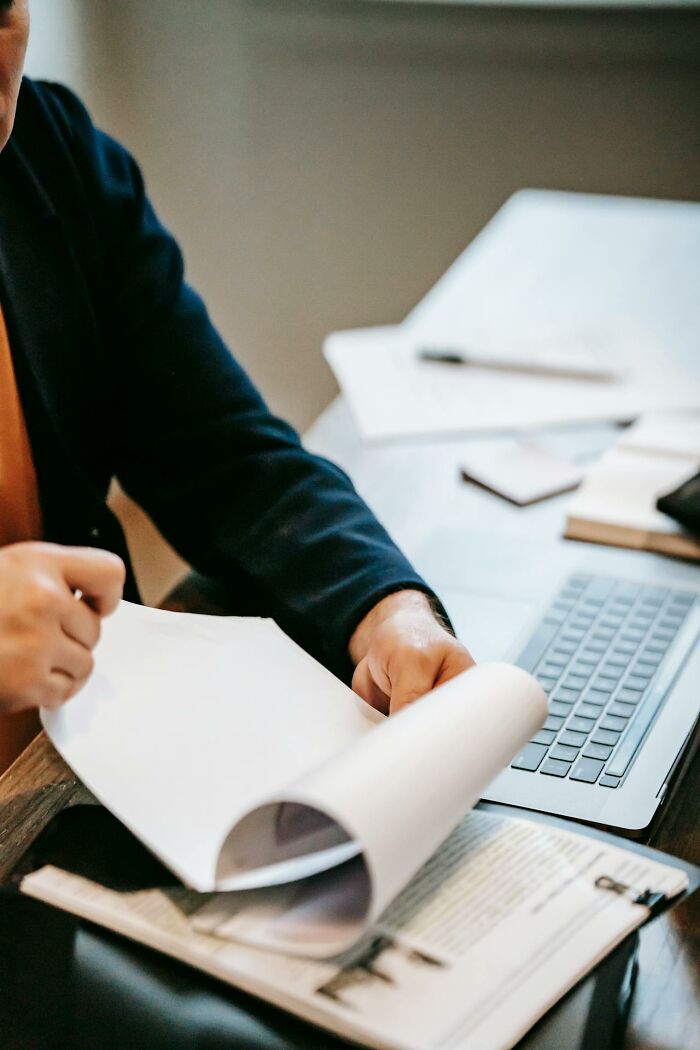 Person flipping through papers at a desk with a laptop, symbolizing power dynamics in a professional setting.