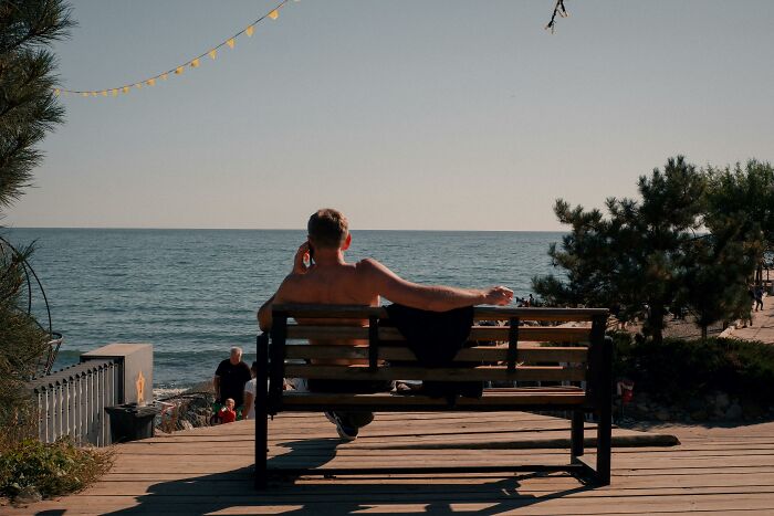 Man sitting shirtless on a bench by the sea, enjoying a peaceful moment associated with rich people lifestyle.