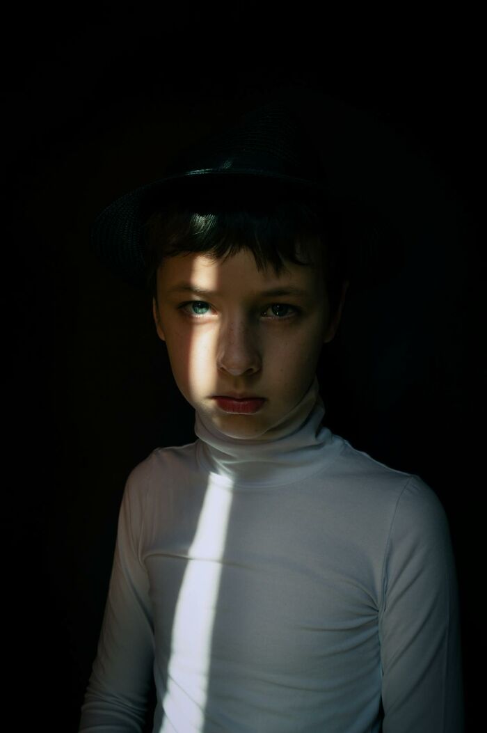 Young boy in a dimly lit room, wearing a hat and white turtleneck, creating a mysterious and creepy atmosphere.