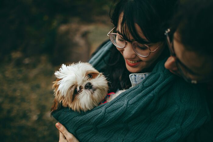 Person smiling and holding a small dog wrapped in a green sweater.